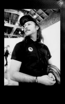 A black and white photo of artist Kurt Brizzolara wearing a black hat and shirt and wearing glasses, from inside the Orange County Center for Contemporary Art gallery.